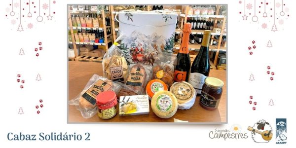 Uma fotografias onde estão vários produtos regionais (uma garrafa de espumante e uma de vinho, vários enchidos, bolos, 3 queijos, doce de abóbora, um enlatado e um frasco de mel) em cima de um balcão. atrás dos produtos está uma caixa de cartão decorada com um Pai Natal dentro da sua carruagem, puxada por duas renas e com montanhas com neve atrás. Ao fundo, na fotografia veem-se vários expositores onde se veem garrafas de rótulos diferentes, embalagens de produtos variados. À volta das fotografias estão vários desenhos natalícios a vermelho (pendentes, árvores de Natal, estrelinhas e patitas) Frase a Azul: Cabaz 2