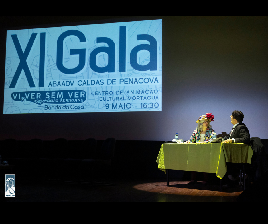 Fotografia onde está um casal num palcos, sentado numa mesa a falar e a olhar para um projeção com texto na parede ao fundo: "XI Gala ABAADV CALDAS DE PENACOVA VIRVER SEM VER Espetáculo às Escuras CENTRO DE ANIMAÇÃO CULTURAL MORTÁGUA com a Banda da Casa 9 MAIO- -16:30 ABAADV"