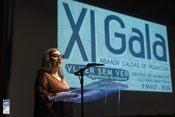 Uma mulher de óculos e com vestido castanho, fala ao microfone. Atrás está uma projeção que diz XI Gala ABAADV Caldas de Penacova Virver sem ver 