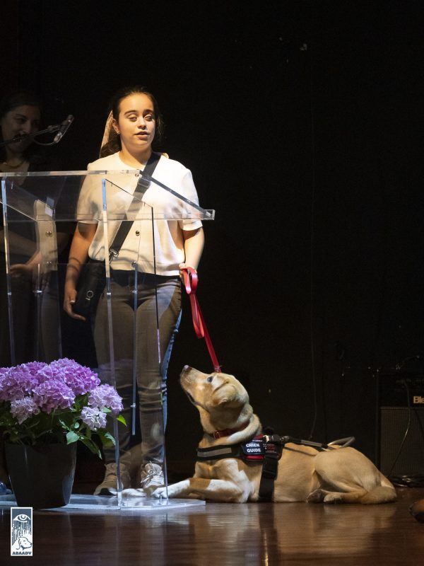 Uma adolescente cega com blusa branca e calças de ganga está num palco a falar num microfone. Está a segurar na trela vermelha de um labrador retriever bege que está deitado ao seu lado e traz um arnês de cão-guia. O cão está a olhar para a jovem. à frente está um arranjo com hortênsias roxas.